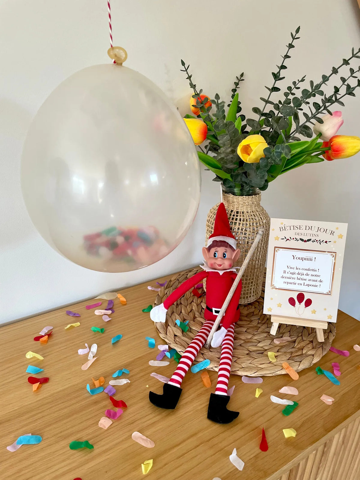 Lutin de Noël assis dans les confettis, bâton à la main, près d'un ballon suspendu et de la pancarte Bêtise du jour